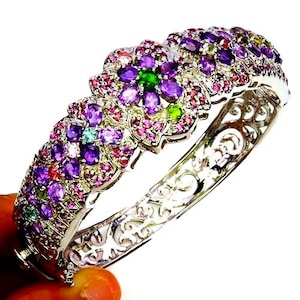 Könnte beinhalten: Ein silbernes Klapparmband mit einem Blumenmuster, das mit oval geschliffenen violetten Amethysten, rosafarbenen Turmalinen und grünen Peridoten besetzt ist. Das Metallband zeigt ein filigranes Durchbruchmuster am unteren Teil.