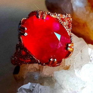 Op de afbeelding: Een ring met een grote, ovaal geslepen, levendig rode edelsteen in een textuurrijke metalen band. De zetting heeft meerdere pootjes die de steen vasthouden, en het metalen oppervlak vertoont een korrelige, onregelmatige afwerking over de hele band.
