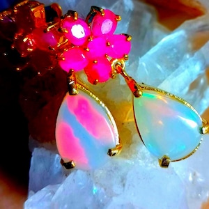 May include: A pair of gold-tone earrings featuring a cluster of vibrant hot pink round stones at the top, connected to dangling, pear-shaped white opalescent stones that display iridescent flashes of pink and blue light within the setting.