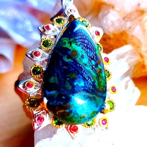 May include: A teardrop-shaped pendant featuring a large, mottled blue and green stone at the centre. The stone is surrounded by a silver setting adorned with alternating small, round, faceted red and green gemstones set in gold-toned bezels.