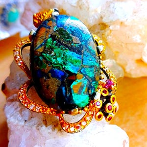 May include: An oval pendant featuring a large, multi-coloured azurite-malachite stone with deep blue, vibrant green, and metallic bronze inclusions. The stone is set in a gold-toned metal frame accented with small white stones and red gems.