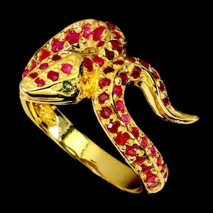 Pode incluir: Um anel de cobra em ouro com um design enrolado, incrustado com numerosos rubis pequenos, redondos e de um vermelho vibrante. A cabeça da cobra é detalhada com uma única pequena pedra preciosa verde-escura para o olho, em metal dourado.