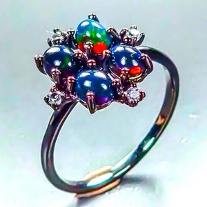 Puede incluir: Un anillo de metal oscuro con un grupo de cuatro ópalos negros redondos e iridiscentes con destellos verdes y rojos vibrantes, acentuado por dos pequeñas piedras blancas redondas y transparentes engastadas en garras sobre una banda fina.