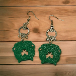 May include: A pair of green glitter pot of gold earrings with a shamrock cut-out. The earrings have gold-tone metal hoops and a dangling charm.