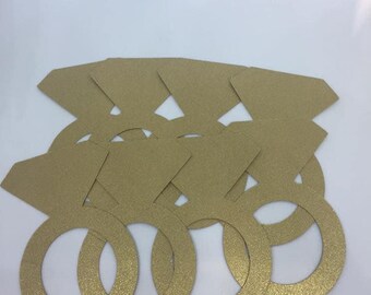 May include: A collection of gold glitter paper ring cutouts with diamond-shaped tops. The rings are arranged in a slightly overlapping pattern, creating a layered effect. The paper has a textured, sparkling finish, and the overall design suggests a wedding or engagement theme.
