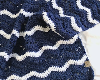 May include: A navy blue and white crocheted blanket with a chevron pattern. The blanket is folded and has a small tag attached to it.