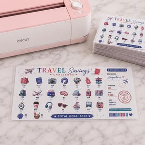 May include: A light pink Cricut cutting machine positioned next to a stack of printed travel savings challenge cards. Each card features a boarding pass design, travel-themed icons with dollar amounts, and the text Travel Savings Challenge.