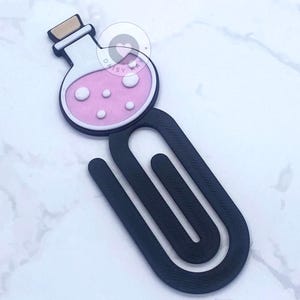 May include: A black paperclip with a potion bottle design. The bottle is white with a pink liquid and white bubbles. The bottle has a brown cork. The paperclip is made of a dark material.