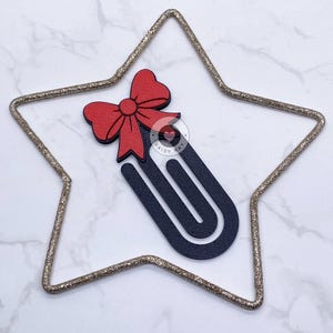 May include: A decorative paperclip with a red bow and a dark blue body, set inside a gold glitter star. The paperclip has the text "DAISY KAY" on it.