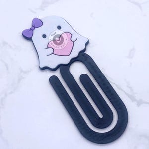 May include: A whimsical ghost-shaped bookmark with a purple bow and a pink heart. The bookmark is white with black accents and a dark gray paper clip design. The text "DAISY KAY" is printed on the heart.