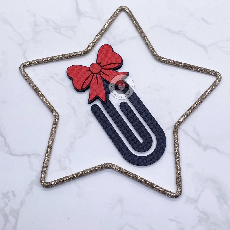May include: A decorative paperclip with a red bow and a dark blue body, set inside a gold glitter star. The paperclip has the text "DAISY KAY" on it.