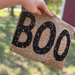 May include: A tan fabric item featuring a leopard print pattern and the word BOO spelled out in large, black, reflective sequins. The letters are arranged horizontally across the center of the fabric, creating a bold, textured visual contrast.