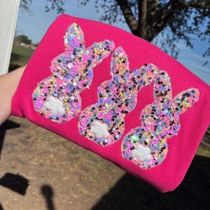 May include: A bright pink sweatshirt featuring three sequined bunny designs. The bunnies are decorated with iridescent sequins in shades of pink, purple, and silver. Each bunny has a white, fluffy tail. The sweatshirt is a festive, Easter-themed garment.