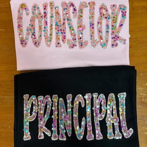 May include: A light pink t-shirt with the word COUNSELOR in multicolored sequin letters and a black t-shirt with the word PRINCIPAL in multicolored sequin letters. Both shirts feature embroidered borders around each individual letter.