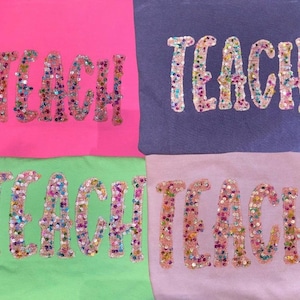 May include: Four fabric shirts in neon pink, purple, light green, and light pink. Each shirt features the word TEACH embroidered in large, block letters filled with multicolored, reflective sequins that create a vibrant, shimmering appearance.