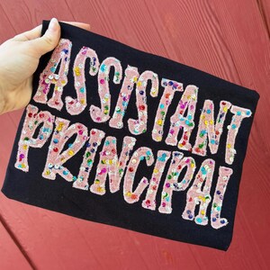 May include: A black fabric item featuring the text ASSISTANT PRINCIPAL in large, light pink, fuzzy, chenille-style letters. The letters are decorated with small, colorful, reflective sequins in shades of blue, green, yellow, and red.