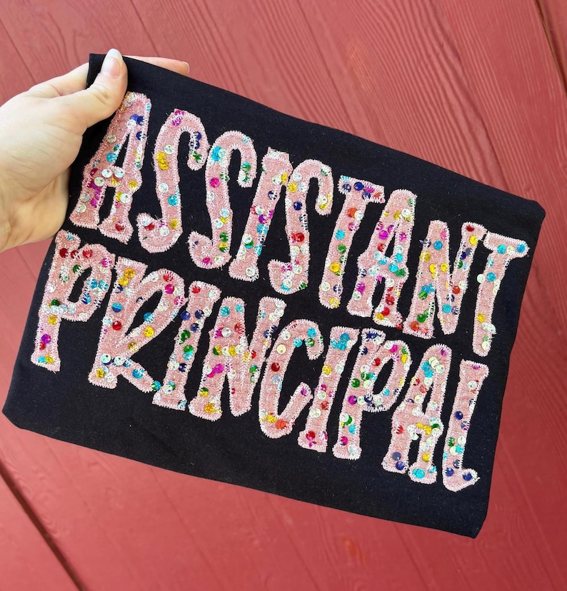 May include: A black fabric item featuring the text ASSISTANT PRINCIPAL in large, light pink, fuzzy, chenille-style letters. The letters are decorated with small, colorful, reflective sequins in shades of blue, green, yellow, and red.