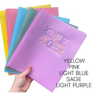 May include: A stack of five translucent plastic folders in yellow, pink, light blue, sage, and light purple. The top light purple folder features the embroidered name KENZIE COOPER in multi-colored thread. Text lists colors: YELLOW, PINK, LIGHT BLUE, SAGE, LIGHT PURPLE.