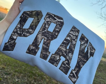 Könnte beinhalten: Hellblaues Sweatshirt mit dem Wort "PRAY" in großen Buchstaben mit Camouflage-Muster. Die Buchstaben sind mit dunklen Nähten umrandet. Das Sweatshirt wird vor einem natürlichen Hintergrund im Freien präsentiert.