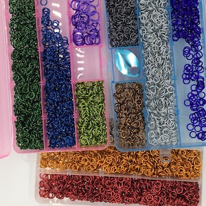 May include: A selection of colourful jump rings in a range of shades, such as blue, pink, green, and gold, arranged in clear plastic containers. These small, metallic rings are suitable for jewellery making and crafts.