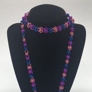 May include: A long necklace made of intertwined metal wire loops in shades of pink, purple, and blue. The necklace is displayed on a black mannequin bust.