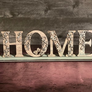 Może przedstawiać: Dekoracyjny drewniany znak z napisem "HOME". Litery są beżowe z misternymi kwiatowymi wycięciami i osadzone na jasnozielonej podstawie. Tło to ciemna, teksturowana powierzchnia.