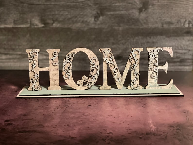 Może przedstawiać: Dekoracyjny drewniany znak z napisem "HOME". Litery są beżowe z misternymi kwiatowymi wycięciami i osadzone na jasnozielonej podstawie. Tło to ciemna, teksturowana powierzchnia.