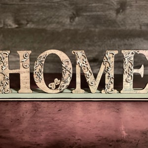 Może przedstawiać: Drewniany znak "HOME" ze skomplikowanymi kwiatowymi wycięciami. Litery są beżowe z wzorem vintage mapy, zamontowane na jasnoniebieskiej podstawie. Dekoracyjny element do wystroju domu.