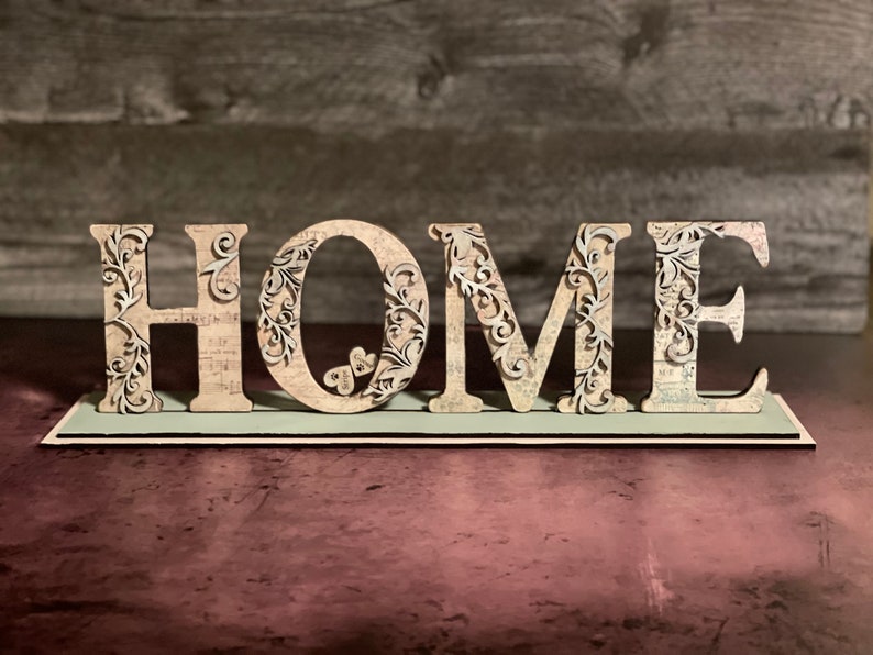 Może przedstawiać: Drewniany znak "HOME" ze skomplikowanymi kwiatowymi wycięciami. Litery są beżowe z wzorem vintage mapy, zamontowane na jasnoniebieskiej podstawie. Dekoracyjny element do wystroju domu.