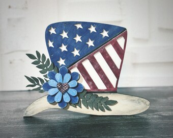 May include: A wooden cutout of a top hat decorated with an American flag design. The hat is adorned with a blue flower with a red heart in the center and green leaves.
