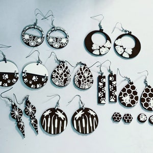 May include: A collection of 14 pairs of wooden earrings with white designs. The designs include flowers, paw prints, honeycombs, dragonflies, and geometric shapes. The earrings are all brown and white.