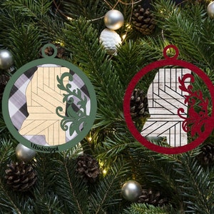 May include: Two round Christmas ornaments, one green and one red, with a white and black checkered background. Each ornament features a cutout of the state of Mississippi with a decorative floral design.