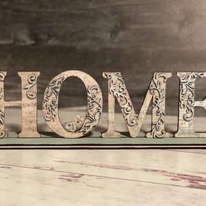Może przedstawiać: Drewniany znak HOME z dekoracyjnym wzorem. Litery są beżowe z misternymi ażurowymi wzorami i kwiatowymi akcentami. Znak stoi na jasnozielonej podstawie, idealny do dekoracji domu.