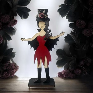 May include: A wooden vampire doll with a red dress, black bat wings, and a black top hat with flowers. The doll is standing on a wooden base.