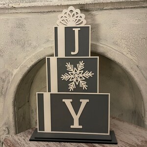 May include: A gray wooden Christmas decoration shaped like stacked boxes with the word "JOY" in white letters. The top box has a white snowflake design and a white decorative bow.
