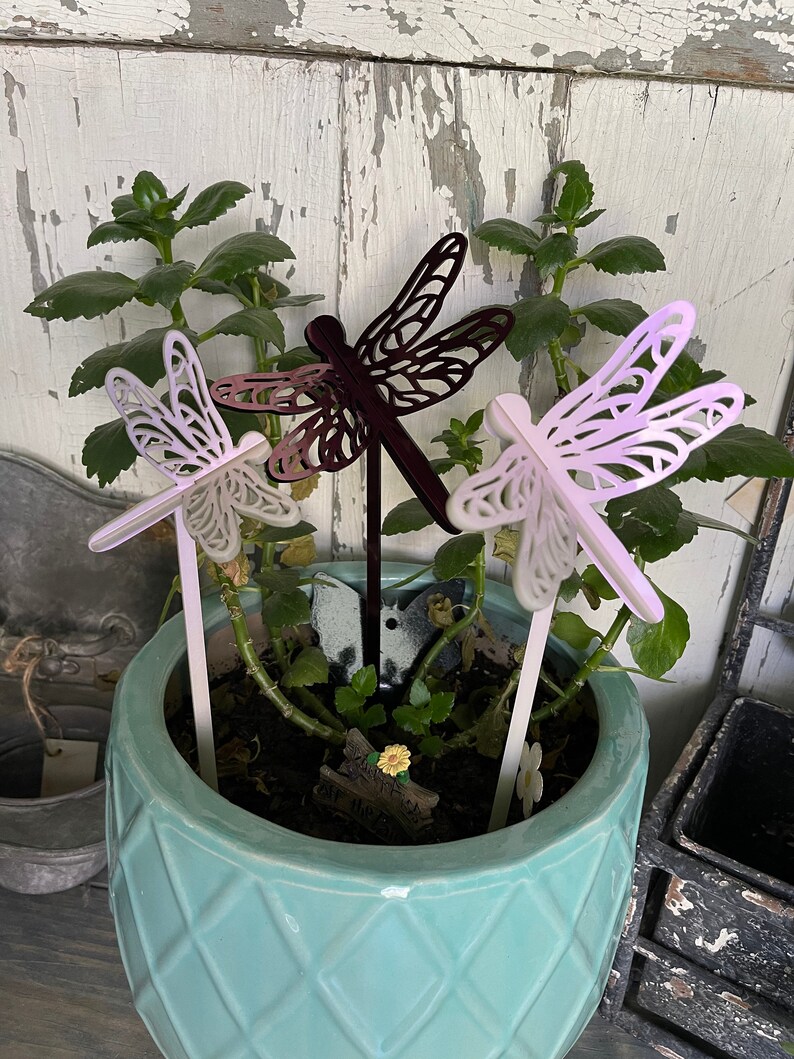 May include: Three metal dragonfly garden stakes in a pot of green plants. The dragonflies are in shades of purple, pink, and black.