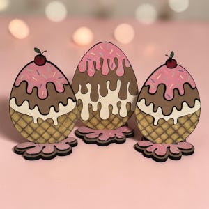 May include: Three decorative wooden Easter egg ornaments shaped like ice cream cones. Each egg features a waffle cone base, chocolate and pink ice cream, sprinkles, and a cherry on top. The ornaments are on a pink surface.