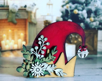 May include: A wooden Christmas elf hat decoration. The hat is red with a gold brim and a small white bell with a red bow. The hat is decorated with a snowflake, leaves, and berries. The background is blurred.
