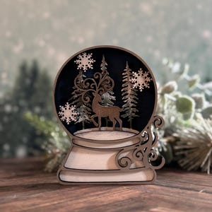 Puede incluir: Decoraci&oacute;n de globo de nieve de madera con un ciervo, copos de nieve y pinos. El globo tiene un interior azul oscuro y una base blanca con remolinos decorativos. El dise&ntilde;o est&aacute; cortado con l&aacute;ser y tiene un tema invernal.