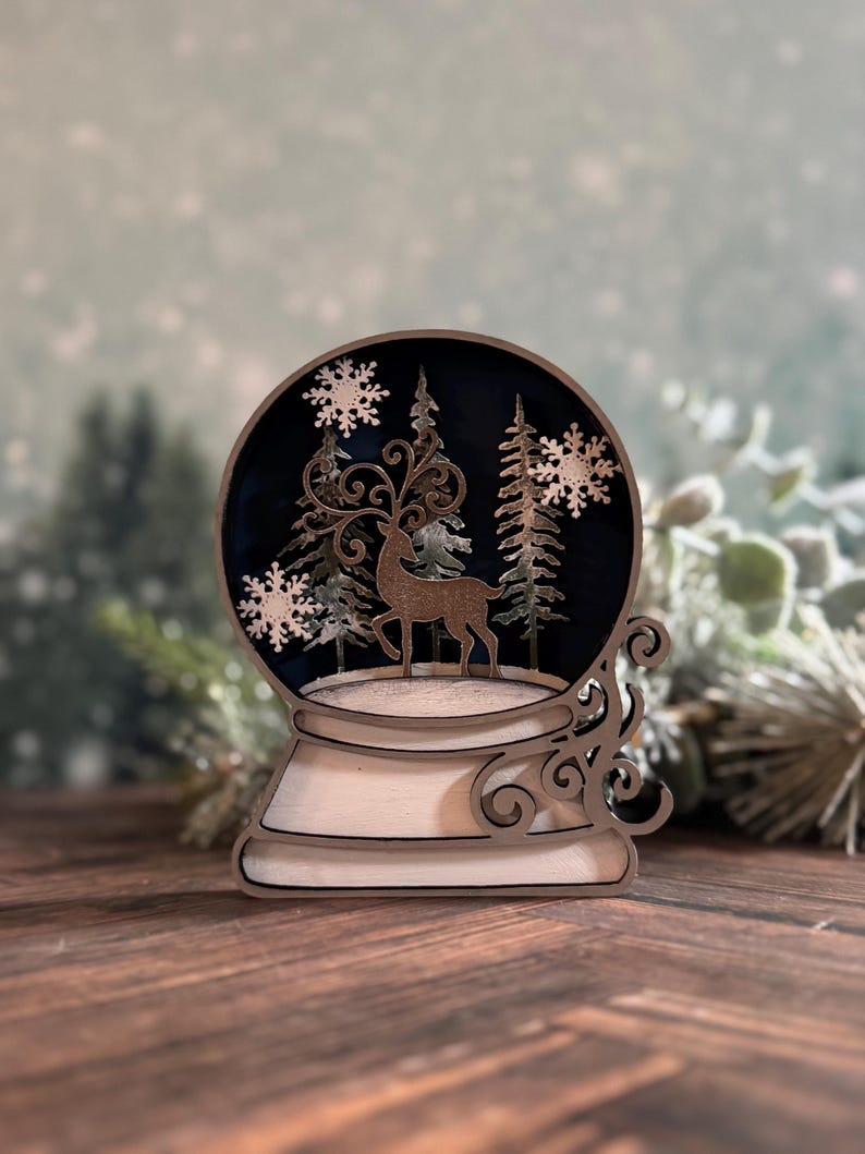 Puede incluir: Decoraci&oacute;n de globo de nieve de madera con un ciervo, copos de nieve y pinos. El globo tiene un interior azul oscuro y una base blanca con remolinos decorativos. El dise&ntilde;o est&aacute; cortado con l&aacute;ser y tiene un tema invernal.
