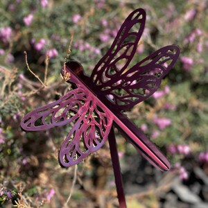 May include: A metal dragonfly garden stake with a burgundy finish. The dragonfly has a detailed, intricate design and is set against a blurred background of purple flowers.