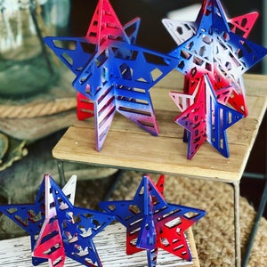 May include: Five wooden stars painted in red, white and blue with a layered design. The stars are arranged on a wooden surface.