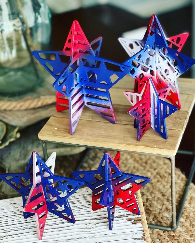 May include: Five wooden stars painted in red, white and blue with a layered design. The stars are arranged on a wooden surface.