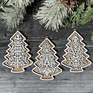Könnte beinhalten: Drei weiße, hölzerne Weihnachtsbaumschmuckstücke mit filigranen, durchbrochenen Designs. Die Schmuckstücke hängen an einem braunen Holzgrund.