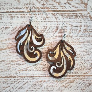 May include: A pair of wooden earrings with a silver-tone finish. The earrings are shaped like a stylized leaf with a swirl design. The earrings are a light brown color.