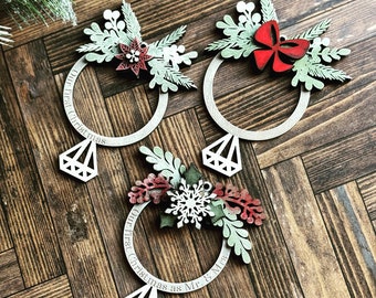 Puede incluir: Tres adornos navideños de madera con forma de anillos con colgantes en forma de diamante. Cada anillo está decorado con vegetación y un lazo o flor roja. El texto "Our first Christmas" o "Our first Christmas as Mr. & Mrs." está inscrito en los anillos.