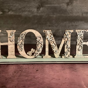 Może przedstawiać: Drewniany znak "HOME" z misternymi kwiatowymi wycięciami. Jasne litery mają wzory kwiatowe i wz&oacute;r vintage na jasnoniebieskiej podstawie. Dekoracja do domu.
