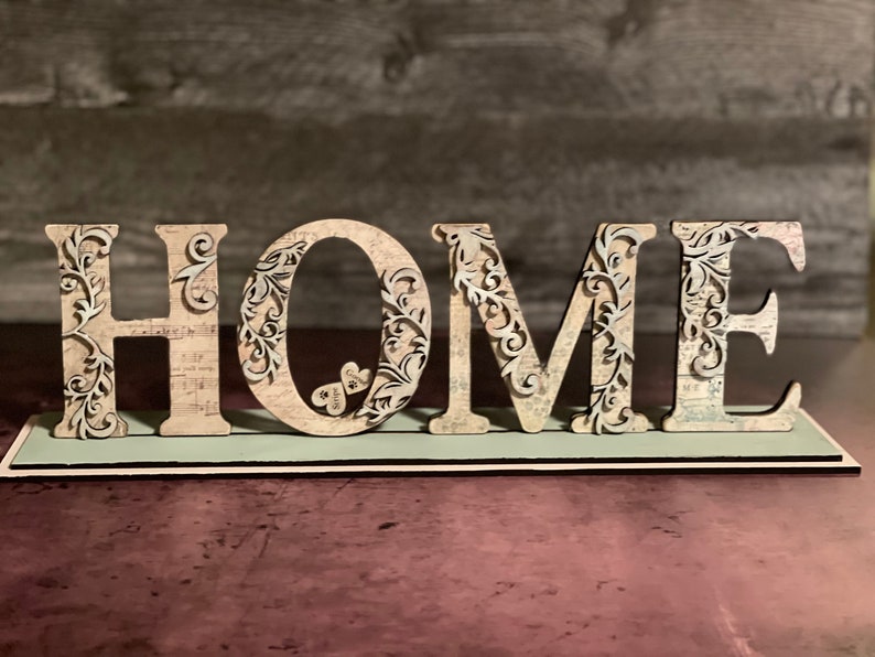 Może przedstawiać: Drewniany znak "HOME" z misternymi kwiatowymi wycięciami. Jasne litery mają wzory kwiatowe i wz&oacute;r vintage na jasnoniebieskiej podstawie. Dekoracja do domu.