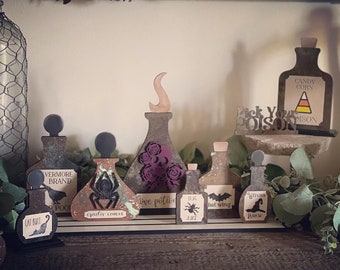 May include: A set of eight Halloween-themed potion bottles with various labels, including "Cat Butt", "Spider Venom", "Love Potion", "Bug Juice", "Bat Wing", and "Witch's Brew". The bottles are made of wood and have a distressed, rustic look. The bottles are arranged on a wooden shelf with a sign that reads "Pick Your Poison".