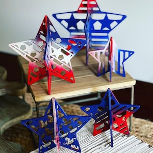 May include: Four wooden stars painted in red, white and blue with a patriotic theme. The stars are different sizes and are standing on a wooden surface.
